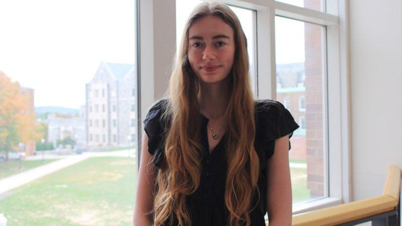 A smiling girl leans against a railing, a big window behind her. 